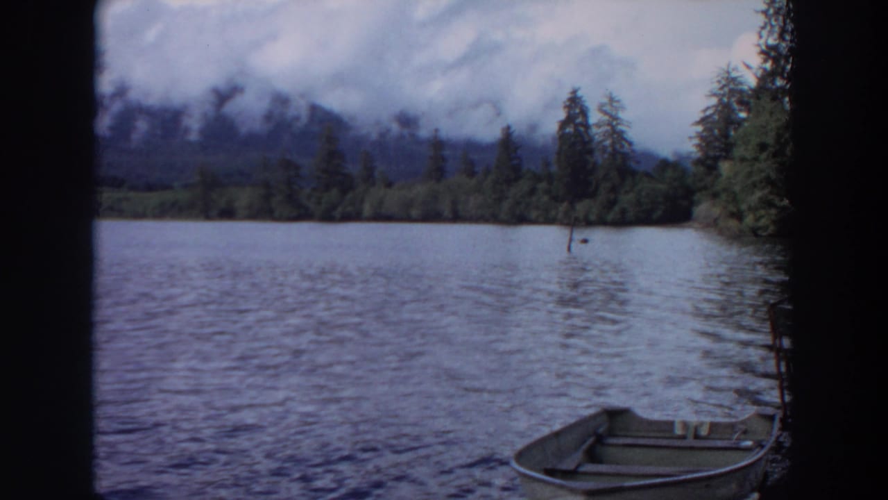 1964 California 8mm view of a calm lake with a small rowboat, evergreen shoreline, cloudy sky, and misty mountains beyond.
