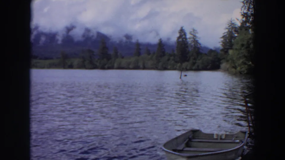 1964 California home-movie frame looking across a rippled lake toward trees and misty mountains, with part of a rowboat in the foreground.