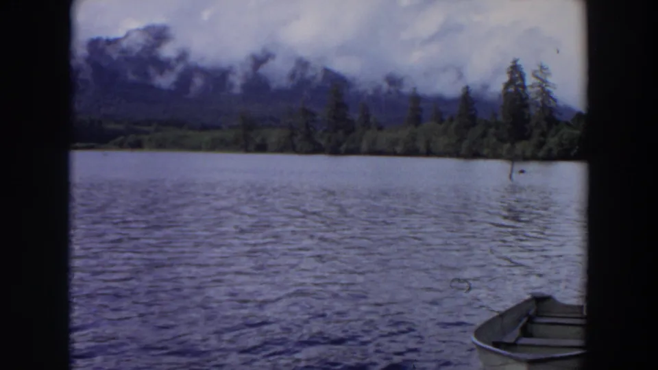 1964 California 8mm frame of a quiet lake, rowboat, forested shoreline, and cloud-covered mountains seen through dark frame borders.