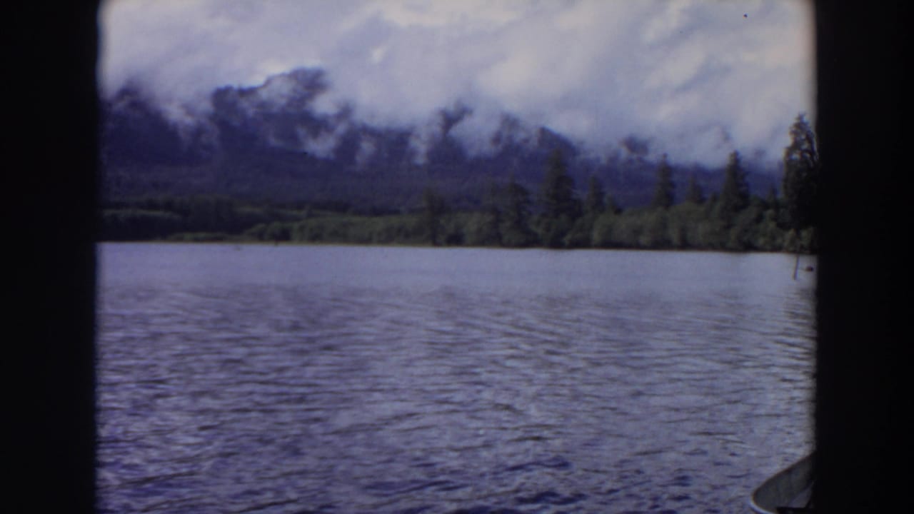 1964 California 8mm frame showing rippled lake water, a forested shoreline, and misty mountains under a cloudy sky.
