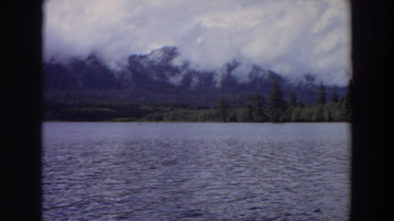 1964 California home-movie frame looking across a lake toward a tree-lined shore and cloud-wrapped mountain ridge.