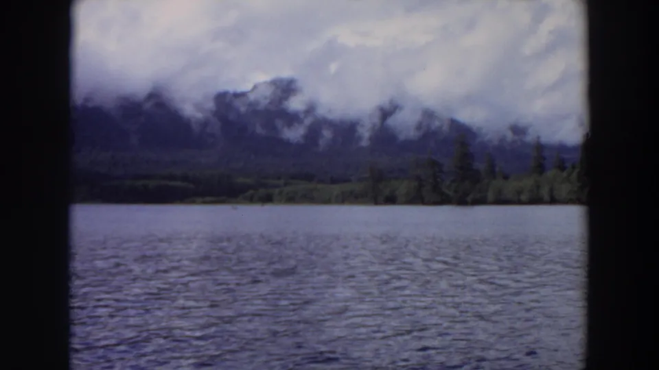 1964 California home-movie frame looking across a lake toward a tree-lined shore and cloud-wrapped mountain ridge.