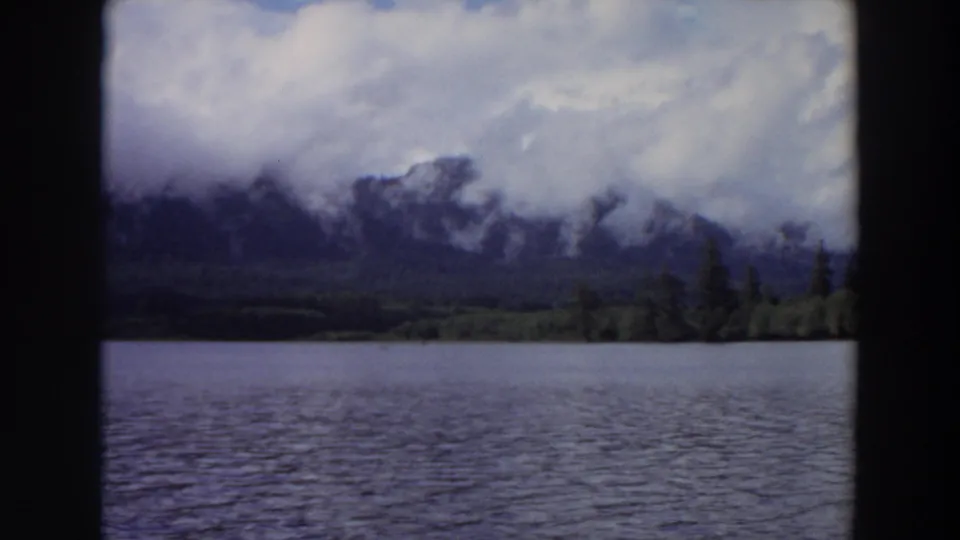 1964 California 8mm still with lake water in front of a forested shore and mountains partly obscured by cloud.