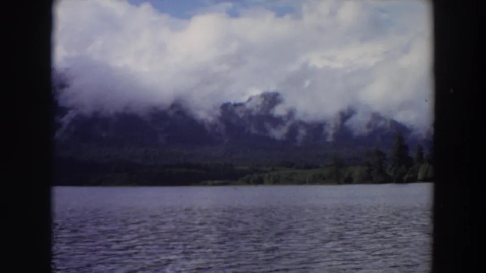 1964 California frame showing a calm lake, tree-covered shore, and misty mountains under a bright cloudy sky.