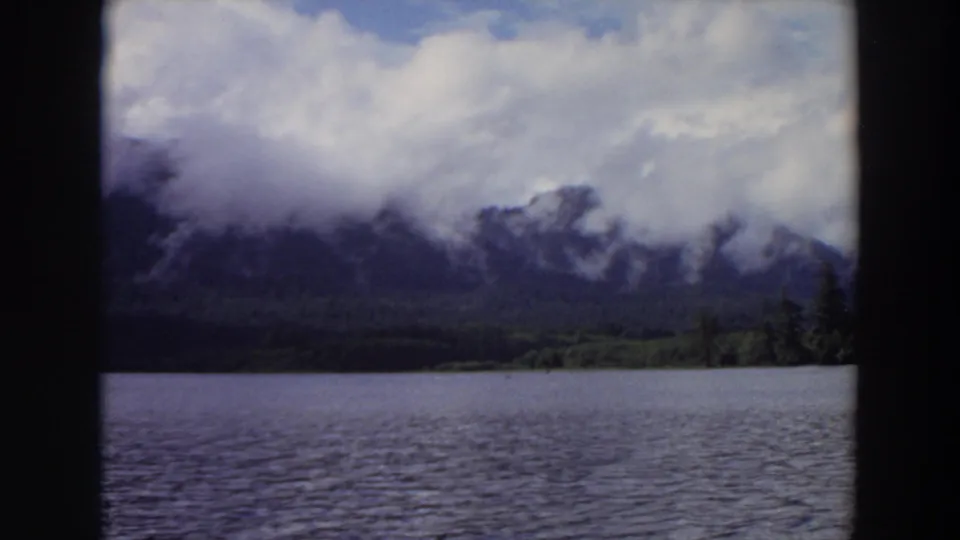 1964 California 8mm frame of a calm lake with dark mountain slopes partly hidden by low clouds.