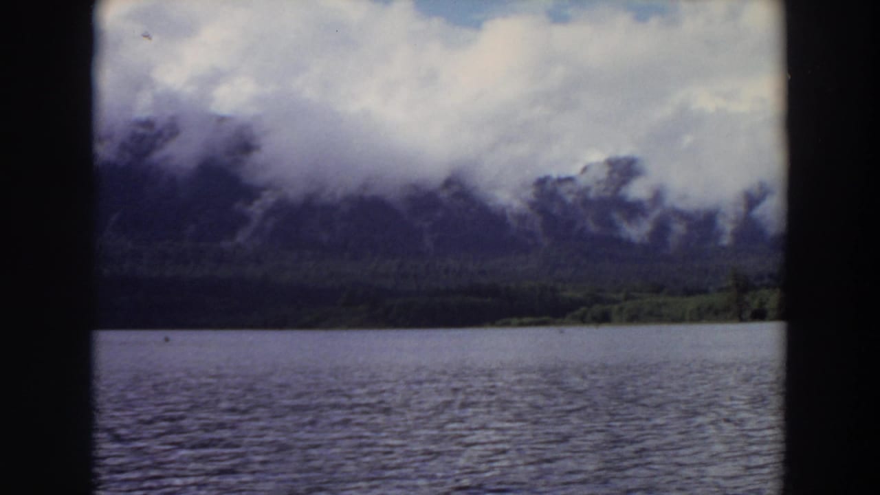 Vintage 8mm view of a California lake, distant forest, and mountains partly obscured by low cloud.