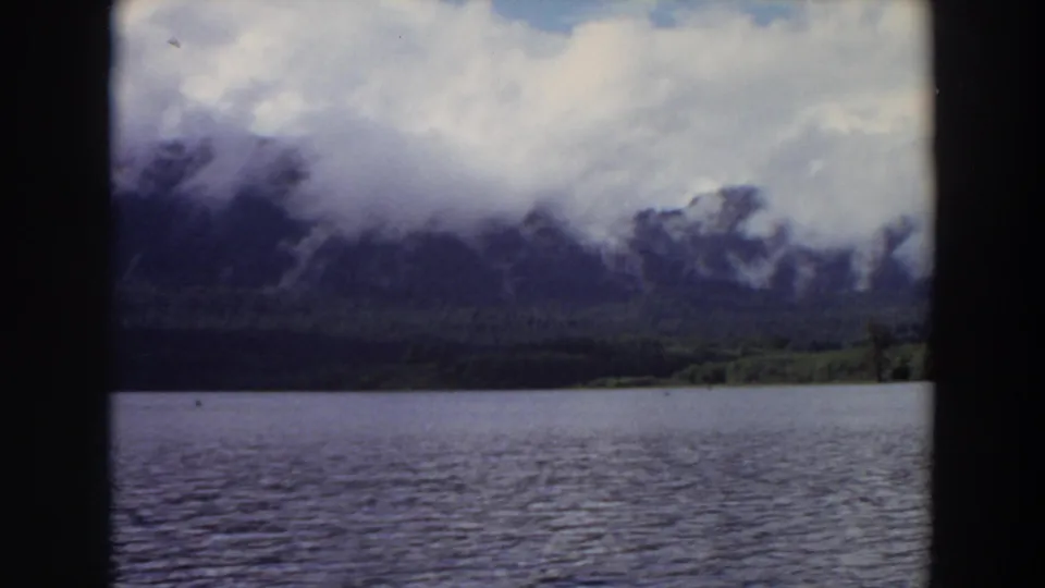 Vintage 8mm view of a California lake, distant forest, and mountains partly obscured by low cloud.
