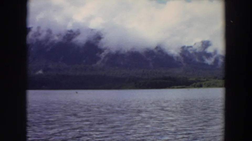 1964 California home-movie frame showing rippled lake water and a cloud-wrapped mountain ridge.