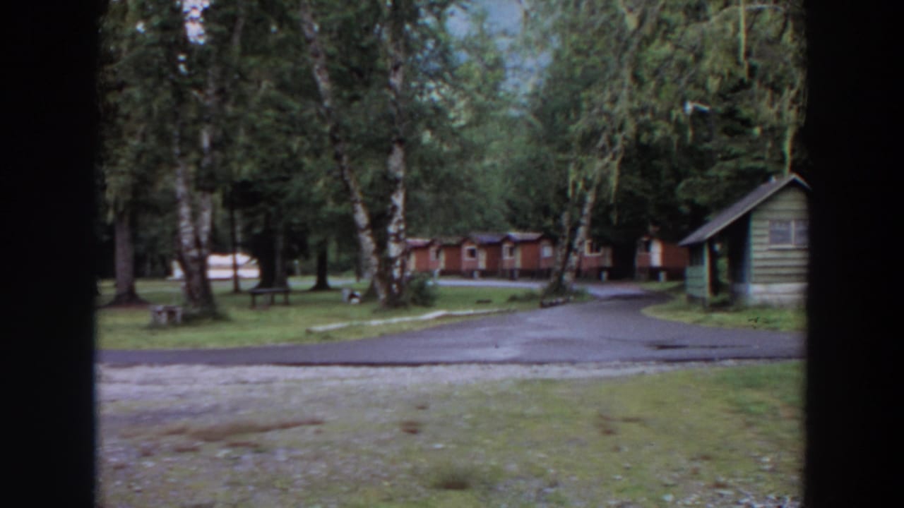 1964 California 8mm frame showing a wooded campground with rustic cabins, green grass, a paved road, and dark borders from a window or film frame.