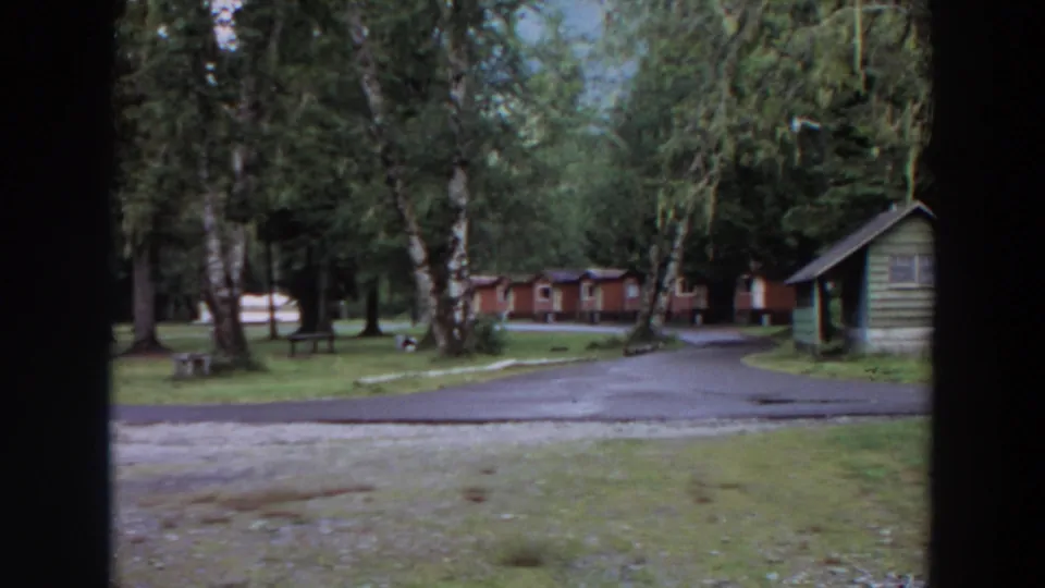 1964 California 8mm frame showing a wooded campground with rustic cabins, green grass, a paved road, and dark borders from a window or film frame.