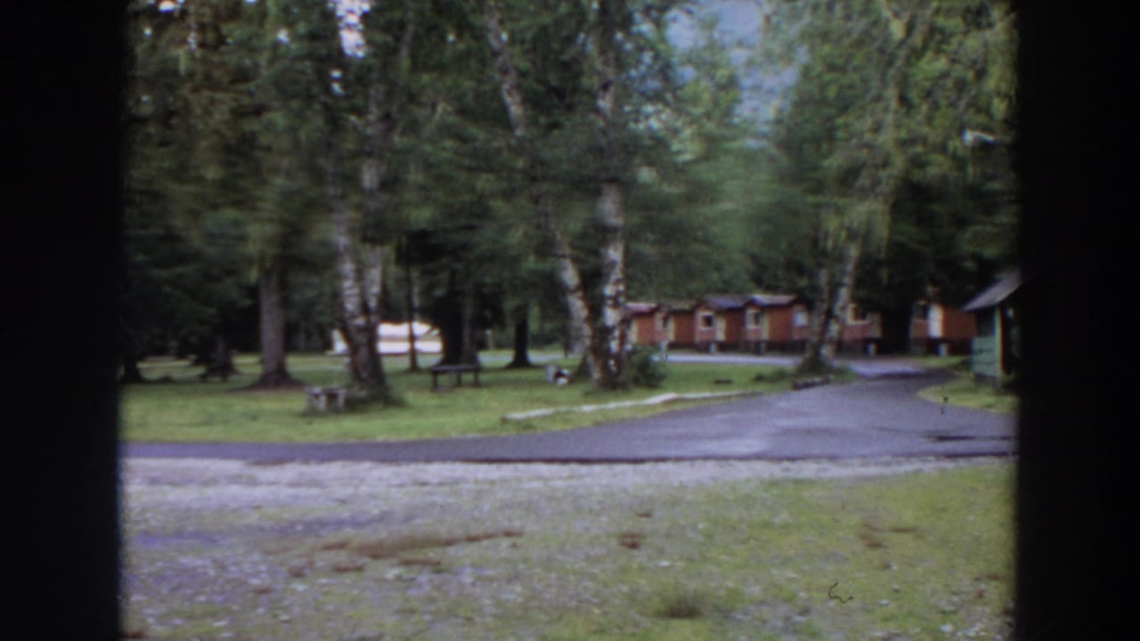 1964 California home-movie frame of a quiet campground road bordered by grass, trees, cabins, and a small structure at the right edge.
