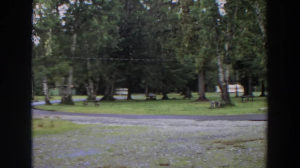1964 California home-movie frame with tall trees, a grassy campground area, a paved road, picnic tables, and cabins partly visible near the right edge.