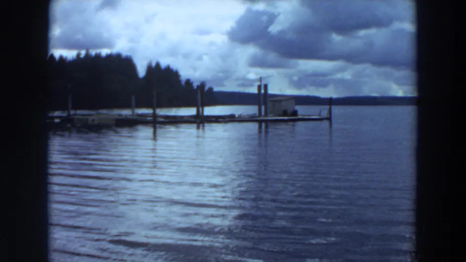 Cloudy lakeside view in California 8mm footage, with dock posts, calm water, distant trees, and a low horizon.