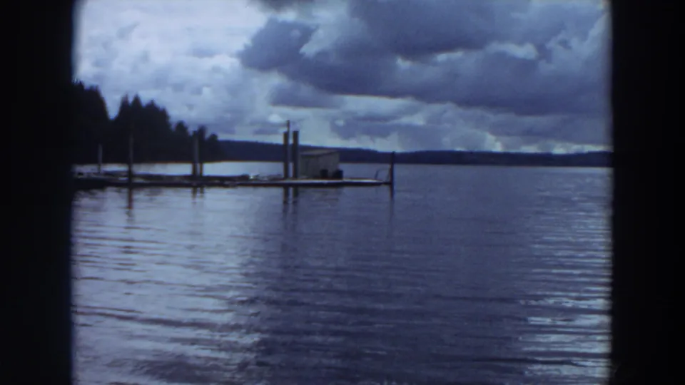 Vintage 8mm lakeside frame showing a dock, reflective water, distant wooded shore, and overcast sky in California.