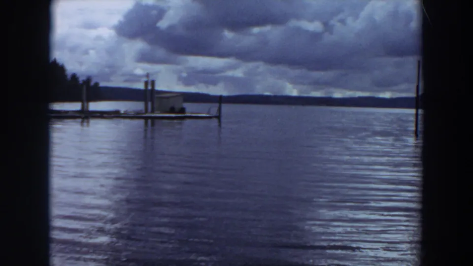1964 California 8mm frame showing dark clouds above a rippled lake, with a distant wooden pier and small lakeside structure.
