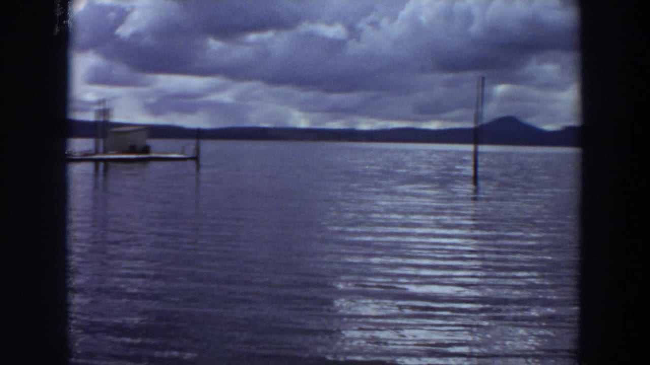 1964 California home-movie frame of open lake water under heavy clouds, with pier posts and a distant hill visible through dark framing.
