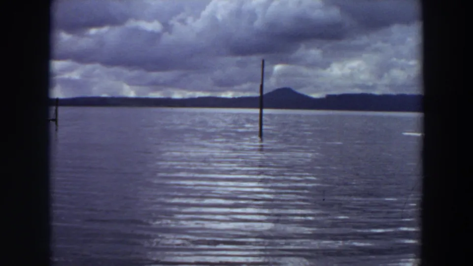 1964 California 8mm view of a single vertical post rising from the lake, backed by storm clouds and distant low mountains.