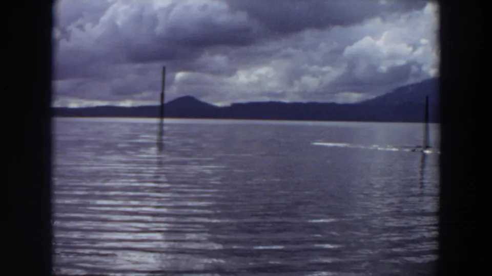 1964 California lake frame showing storm clouds, a vertical post, distant hills, and a small sailboat near the right edge.