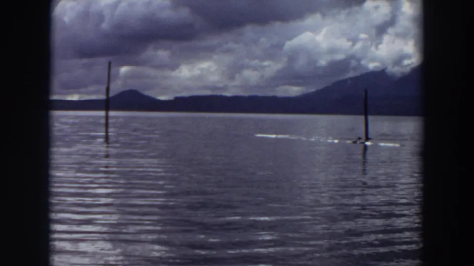 1964 California 8mm frame showing calm lake water, distant mountains, dramatic clouds, and two vertical poles rising from the water.