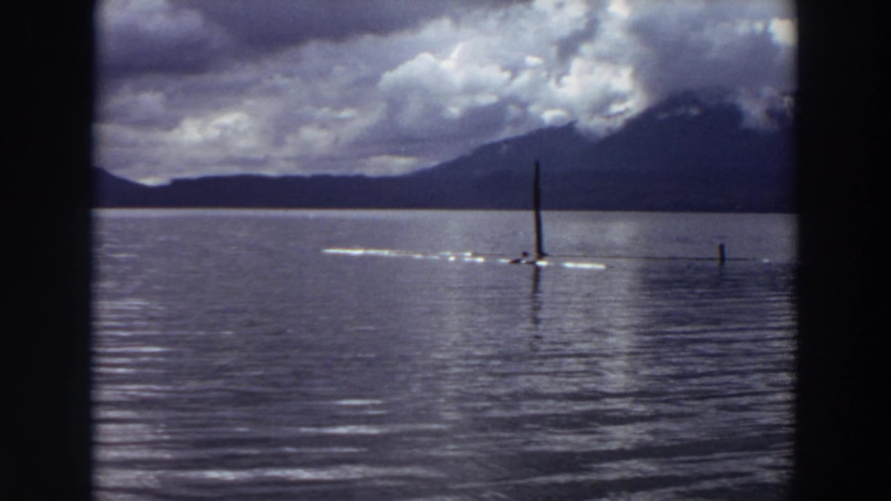 1964 California 8mm frame of a small craft crossing calm lake water near upright poles, with mountains and clouds behind it.