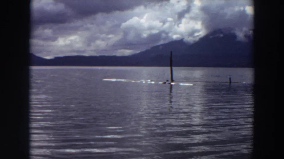 1964 California 8mm frame of a small craft crossing calm lake water near upright poles, with mountains and clouds behind it.