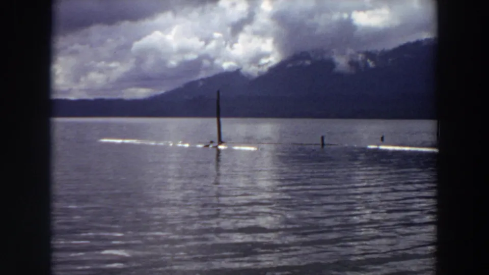 1964 California 8mm frame showing a wider lake view with a bright wake across the water, vertical poles, mountains, and heavy clouds.