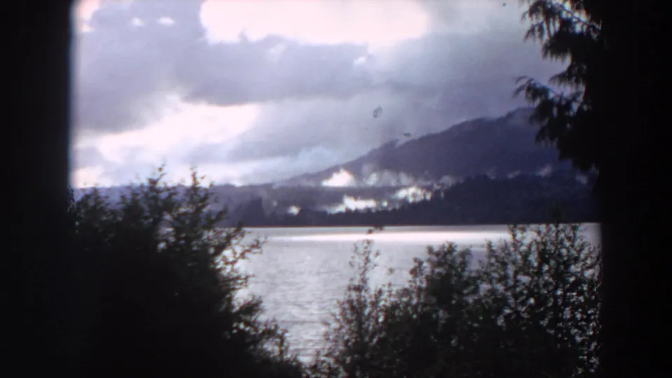 1964 California 8mm frame showing calm water, misty distant mountains, cloudy sky, and dark foreground shrubs.