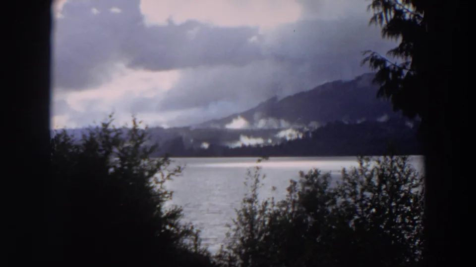 1964 California 8mm view of a mountain lake framed by dark foreground trees, with distant peaks under low clouds.