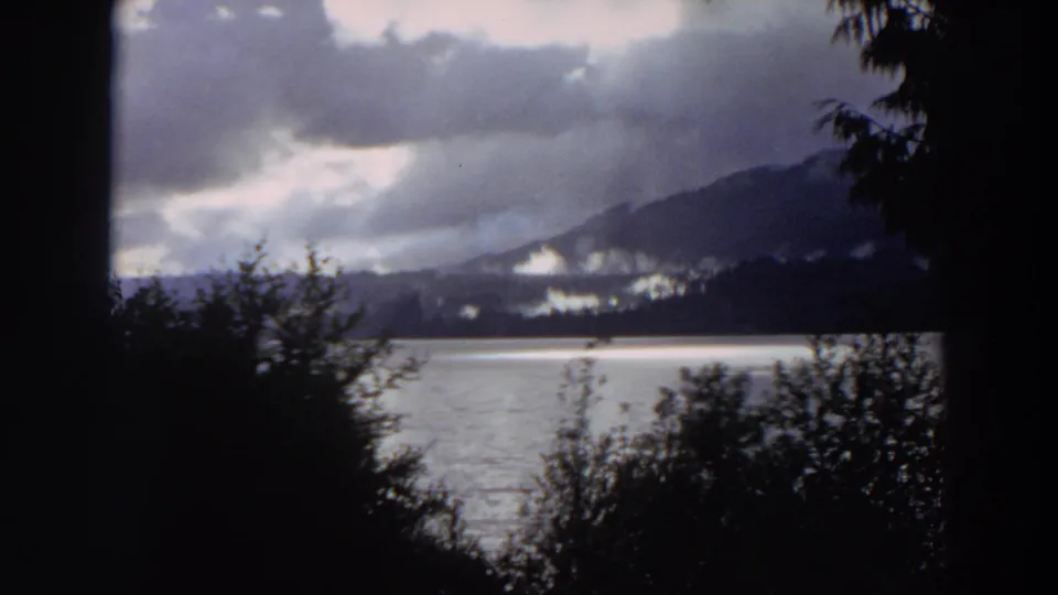 1964 California 8mm frame showing calm lake water, dark trees, and cloud-covered mountain ridges beyond the shore.