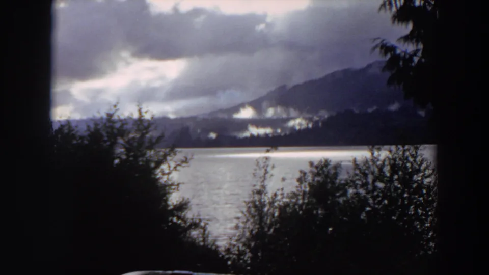 1964 California home-movie still of a lake and mountain ridge, partly obscured by dark shrubs and trees in the foreground.