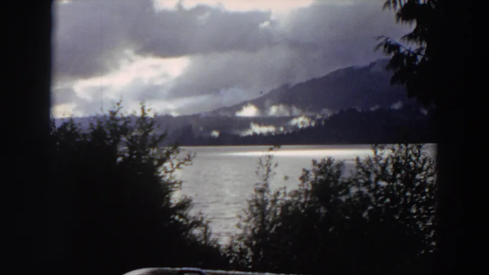 1964 California 8mm view of a mountain lake with dark trees in front, pale water, and cloudy peaks in the distance.