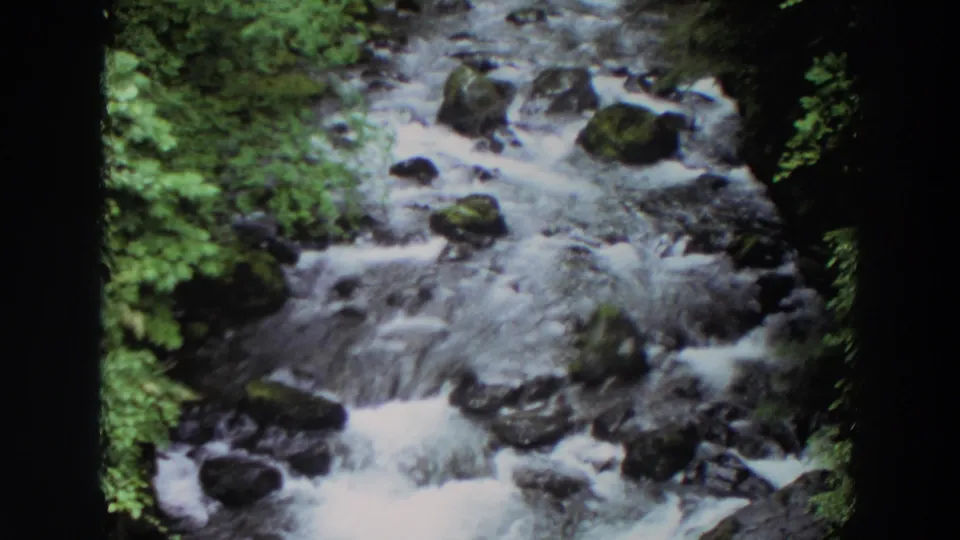 1964 Washington 8mm frame showing the creek from a slightly wider angle, with water moving around rocks and green banks.