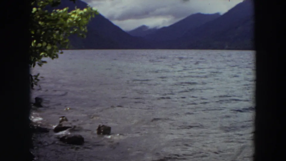 1964 Washington 8mm frame of rippling water and distant mountains, with rocks and foliage marking the shoreline viewpoint.