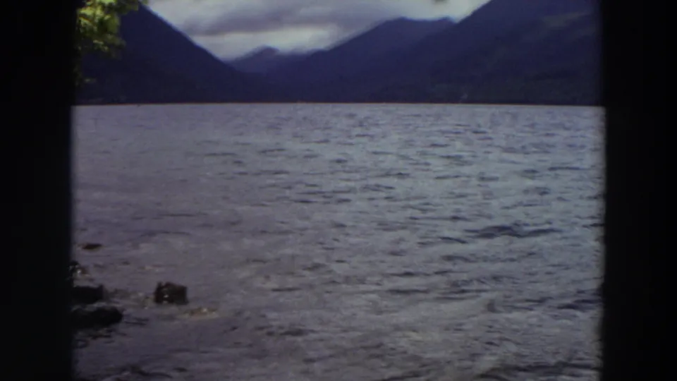 1964 Washington 8mm frame showing a wide stretch of rippling water beneath distant mountains, framed by dark foreground tree edges.