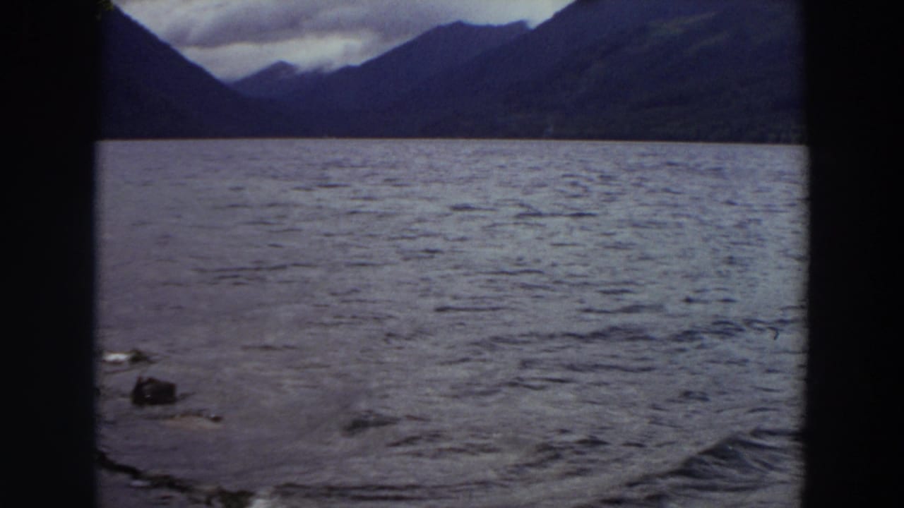 1964 Washington home-movie view of rippled lake water, dark mountain slopes, and overcast sky framed by black film edges.