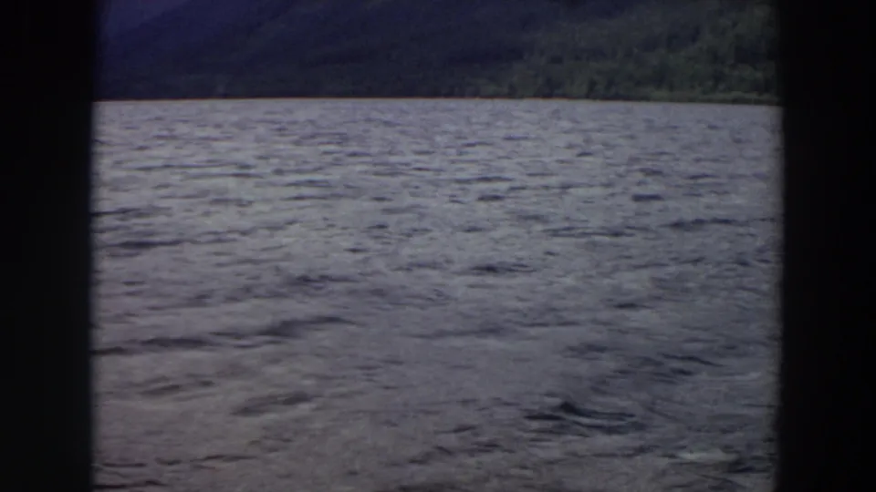 1964 Washington home-movie frame showing rippled lake water, a low forested shore, dark mountain slope, and soft faded film color.