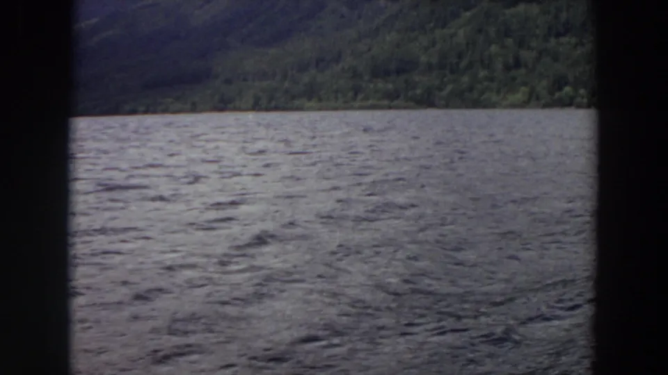 1964 Washington home-movie still of rippled lake water under cloudy light, with a dense green shoreline beyond the dark frame edge.