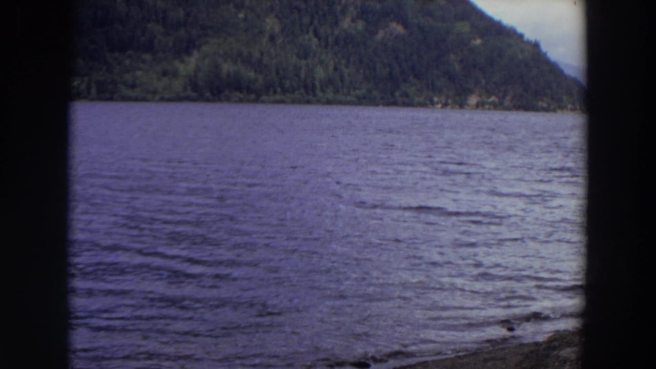 1964 Washington 8mm frame showing rippling mountain water, a forested slope, cloudy sky, and dark edges from the filming position.