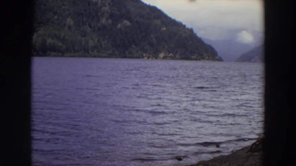 1964 Washington home-movie frame looking across rippled water toward dark wooded mountains and a cloudy valley opening.