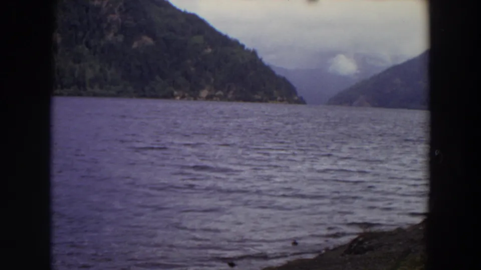1964 Washington 8mm view across purple-tinged water toward wooded slopes, a distant valley, low clouds, and rocky foreground shore.