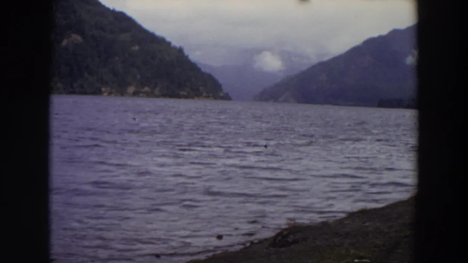 1964 Washington frame showing a wide lake or inlet surface, wooded mountain slopes, a cloudy gap, and rocky shore in the foreground.