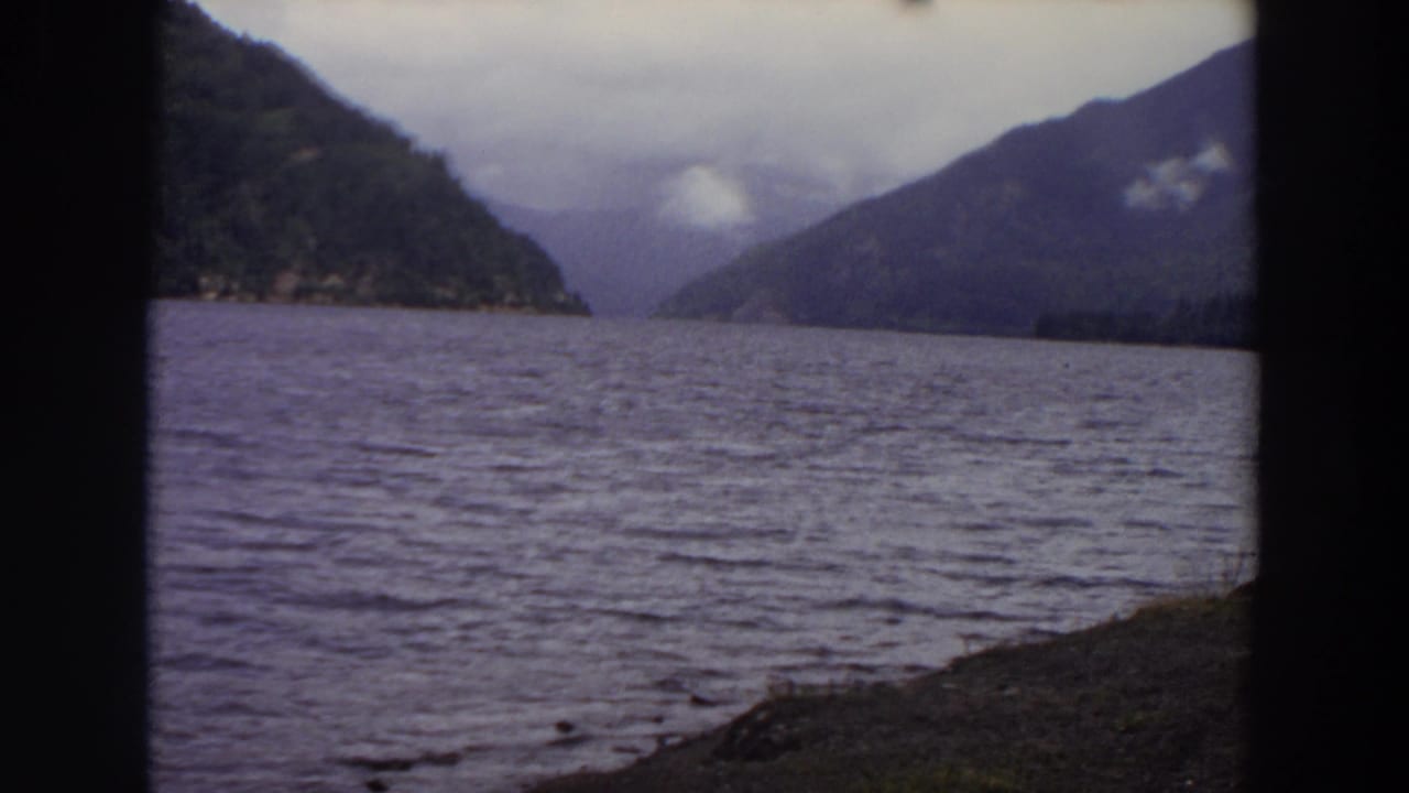 1964 Washington 8mm view of a calm lake between forested mountains, seen through dark vertical edges with a cloudy sky above.