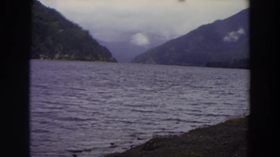 1964 Washington 8mm view of a calm lake between forested mountains, seen through dark vertical edges with a cloudy sky above.