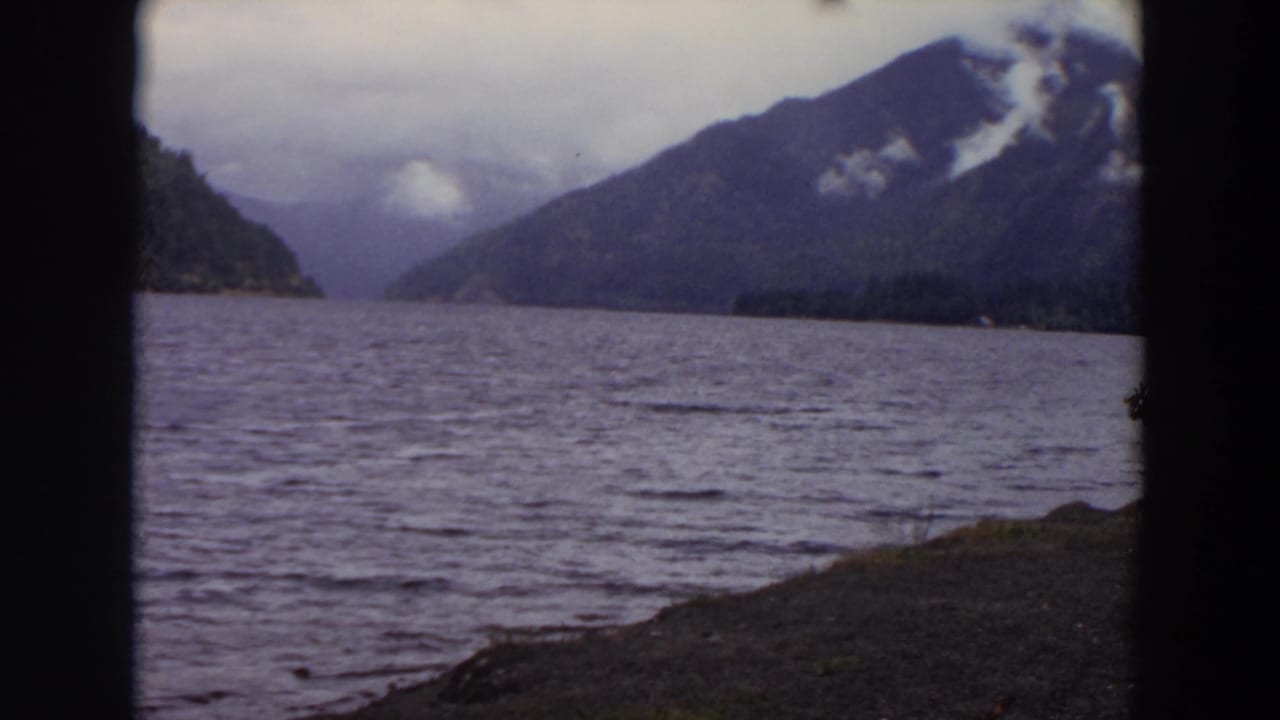 Mountain lake under clouds with a small wooded landform and dark edge framing.