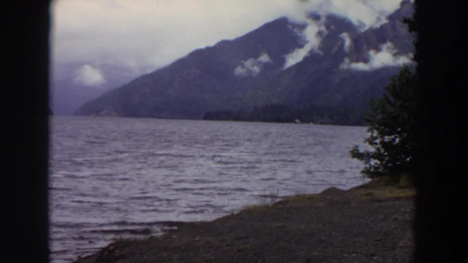 1964 Washington lake scenery with a forested mountain, low clouds, rippled water, a gravel shore, and a tree edge entering the frame.