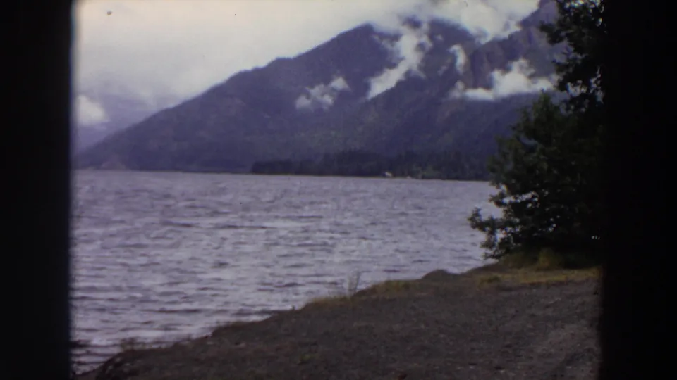 Final 1964 Washington 8mm frame of a mountain lake, with low clouds on the forested slope and shoreline vegetation at the right edge.