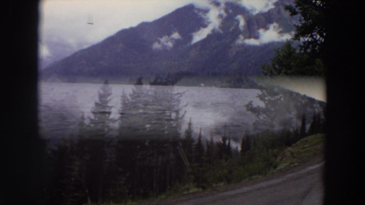 Mountain lake seen through dark frame edges with cloudy peaks, forested shore, and a roadside foreground.