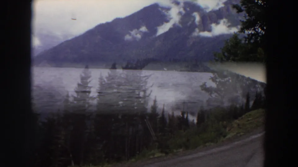 1964 Washington 8mm view of a calm mountain lake, evergreen slopes, low clouds, and a road or gravel edge framed by dark borders.