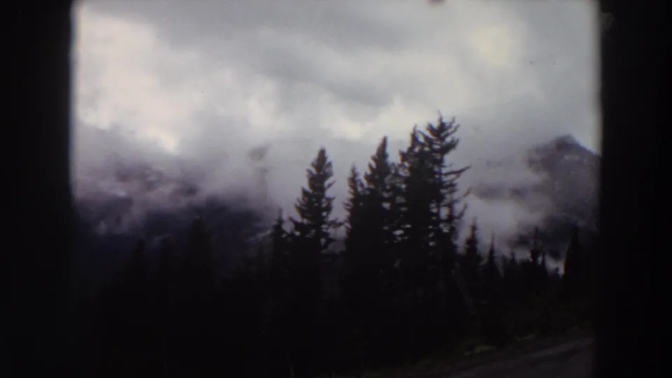 1964 Washington mountain view with heavy cloud cover, evergreen silhouettes, and dark frame edges from the home-movie camera position.