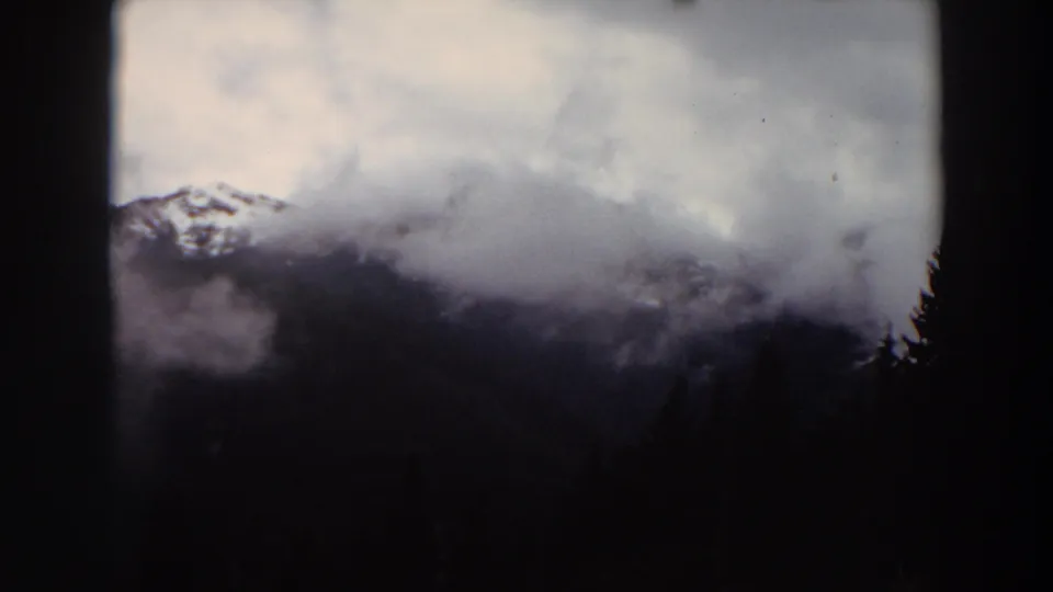 1964 Washington 8mm frame showing a snow-dusted mountain peak partly veiled by clouds over a dark evergreen forest.
